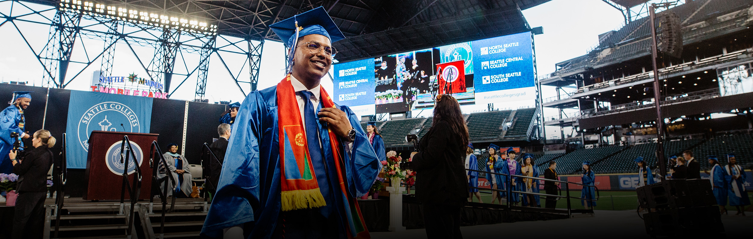 a smiling student in a cap and gown walking off the commencement stage at T-Mobile Park