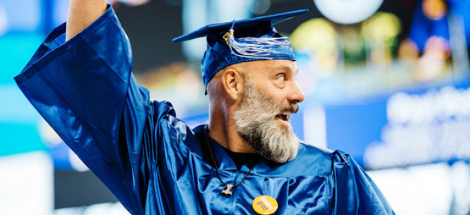 A male graduate with a beard who is smiling and waving