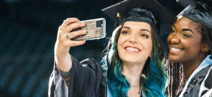 two graduates in caps and gowns taking a selfie with their phones