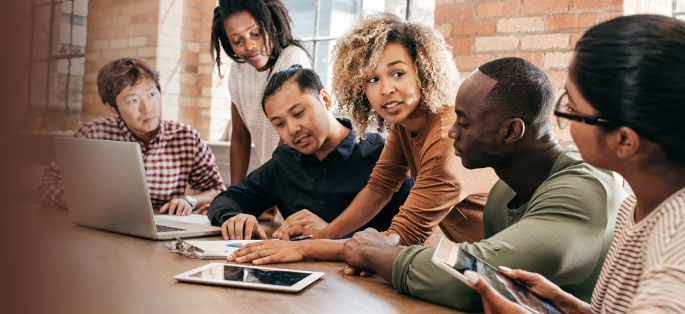 a diverse team of people talking at a table with computers