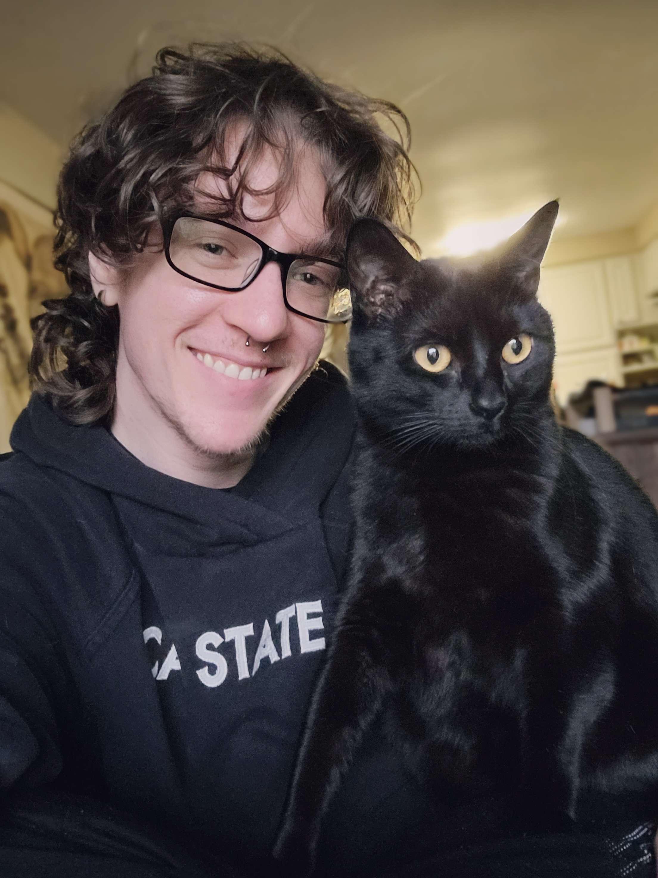 Eli wearing glasses looking at camera with his black cat, Judie next to him also looking at the camera.