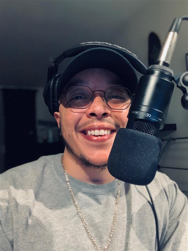 Jesse in recording studio wearing a hat and glass, smiling with grey shirt and a microphone in front of his face.