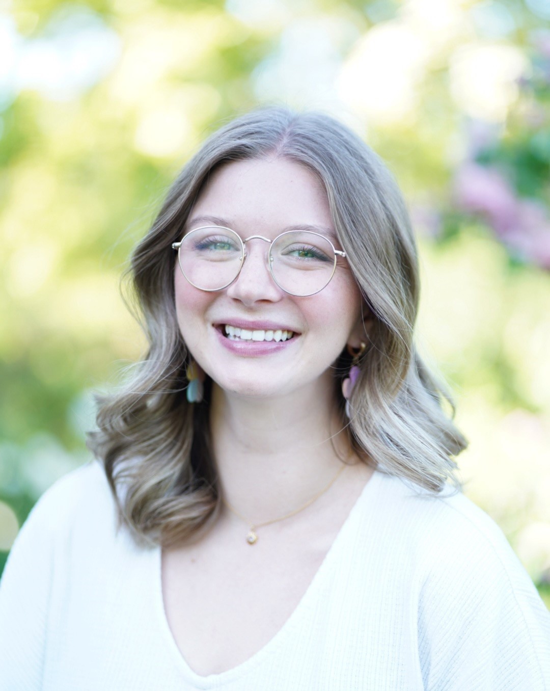 Jessica Standing outside with blurred foliage in the background, smiling with glasses and white shirt.