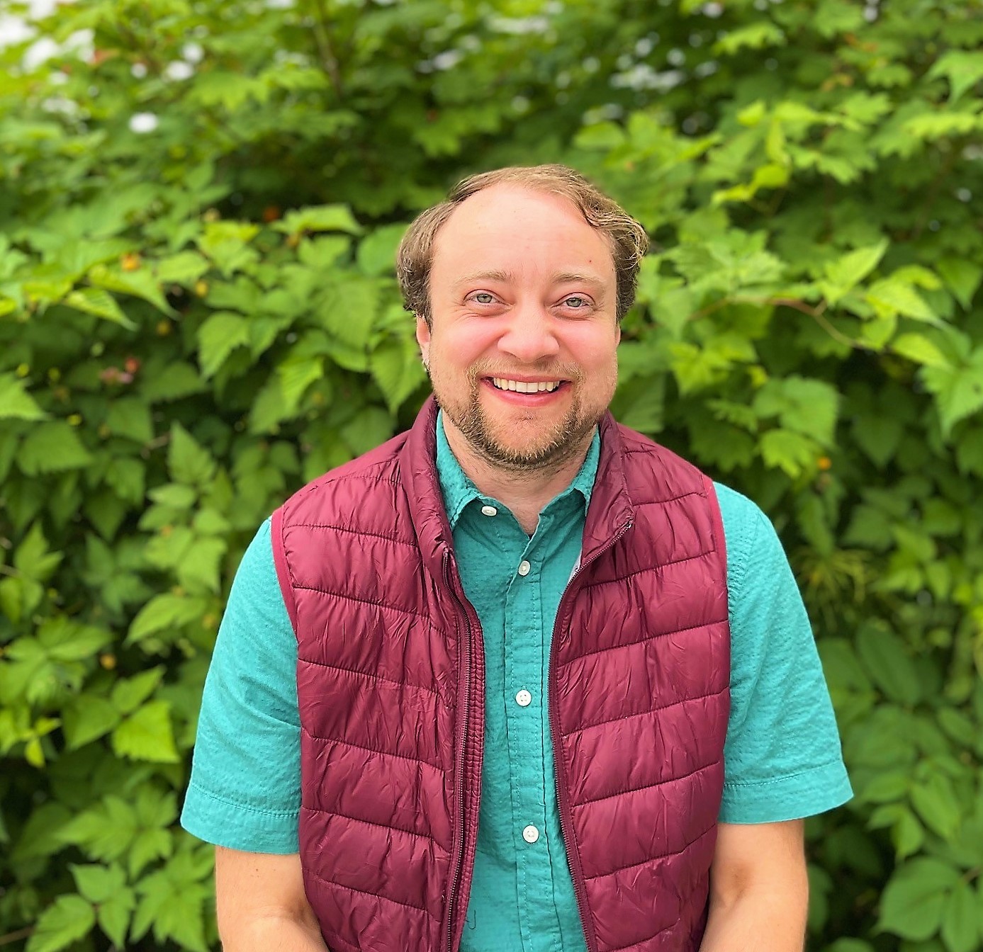 Julius outside with green foliage behind him, smiling with green shirt and red vest.