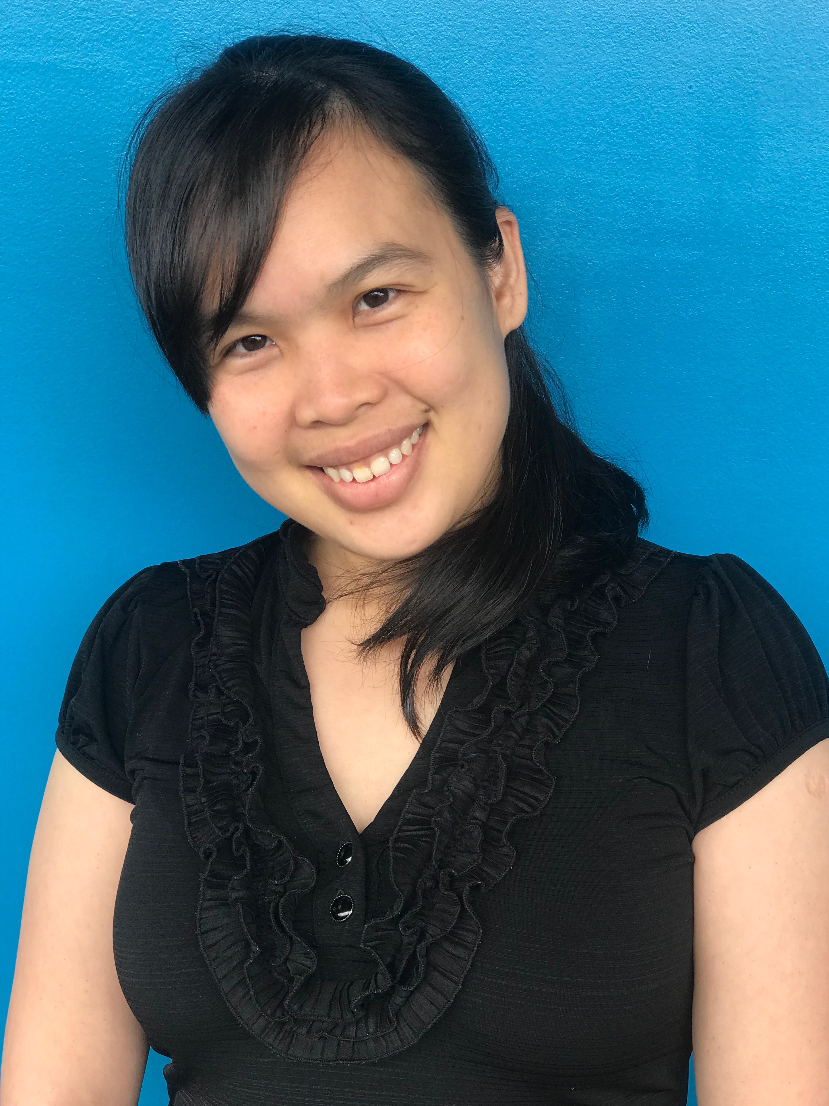 Thuy standing with blue background head slightly tilted, long hair, smiling looking at the camera.